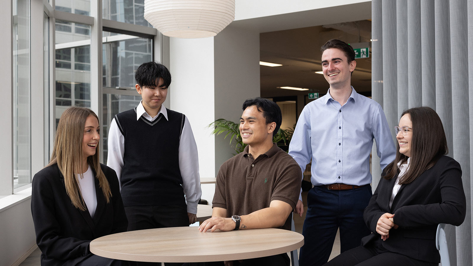 Five RACV graduates smiling together around a table. Three graduates are sitting and two graduates are standing.