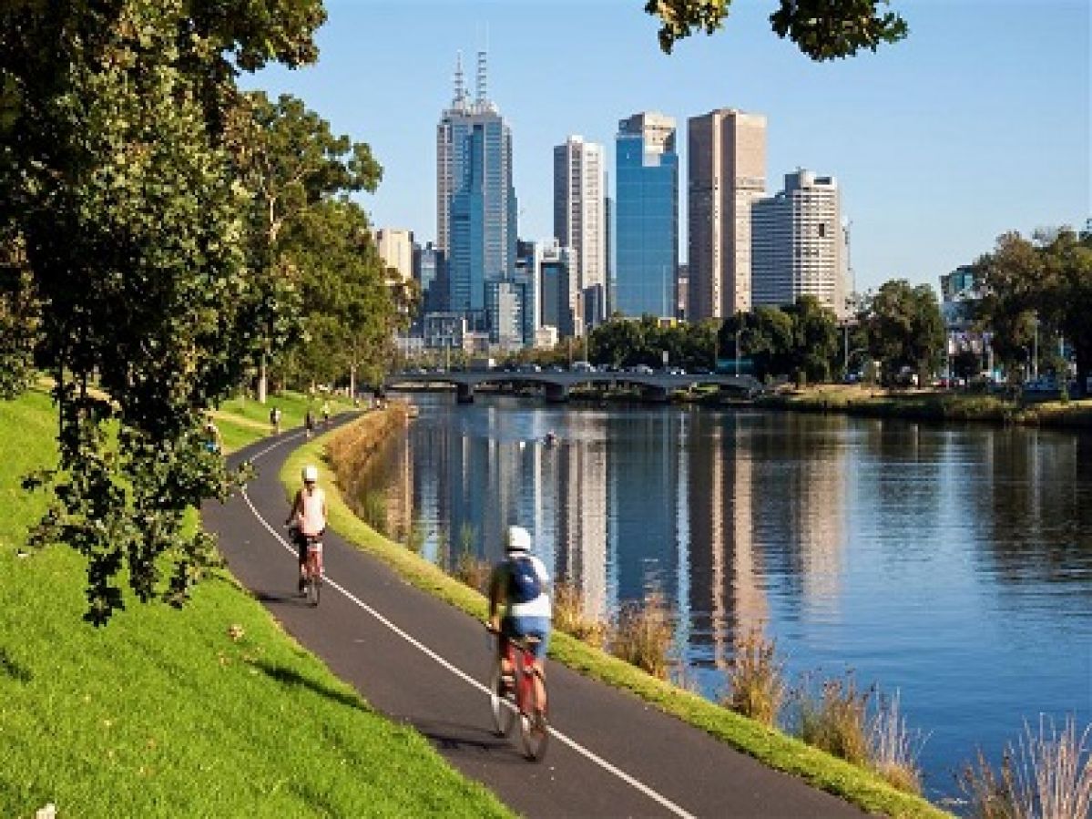 Two people riding a bike along the Yarra River in Melbourne with the city skyline in the background.
