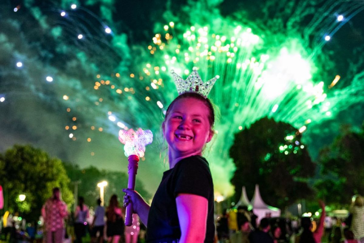 A young girl smiles while carrying a light up wand while a burst of green fireworks explodes in the sky behind her