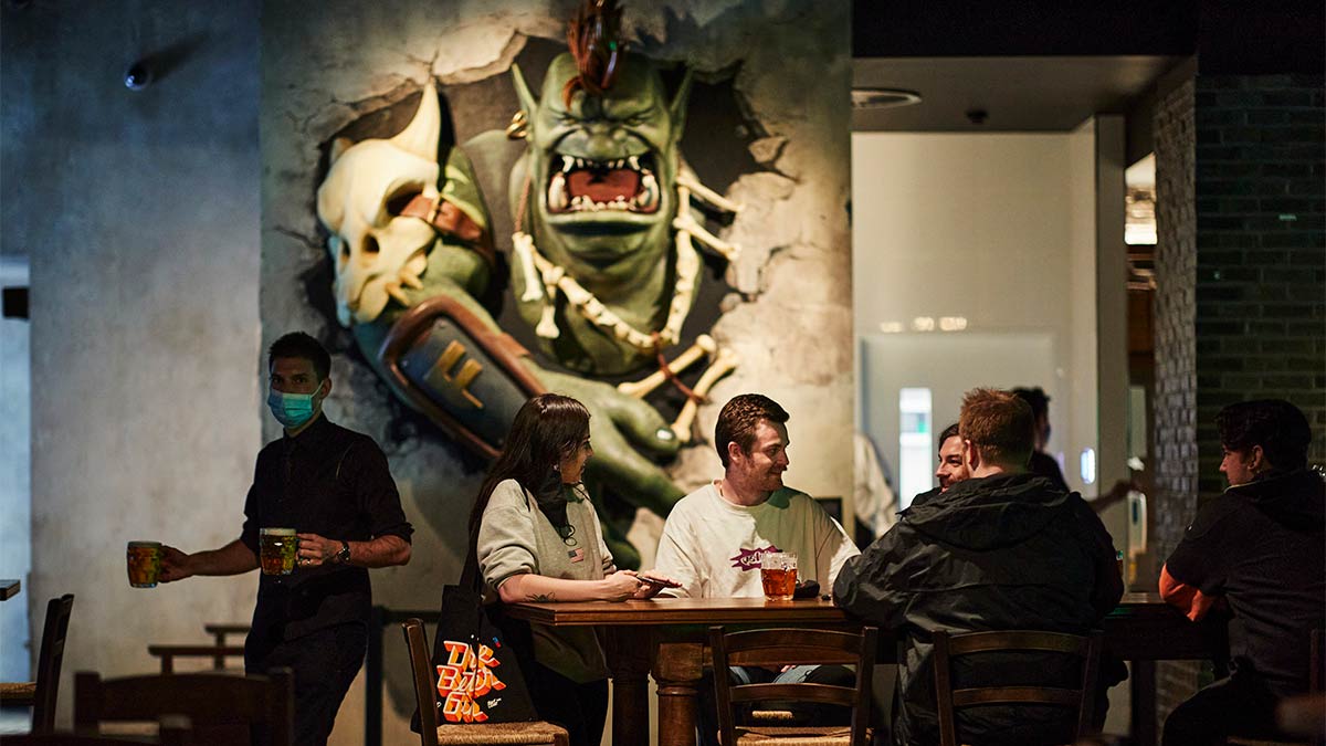 A group of people enjoy beers at a table while a huge sculpture of an ogre bursting through the wall is behind them.
