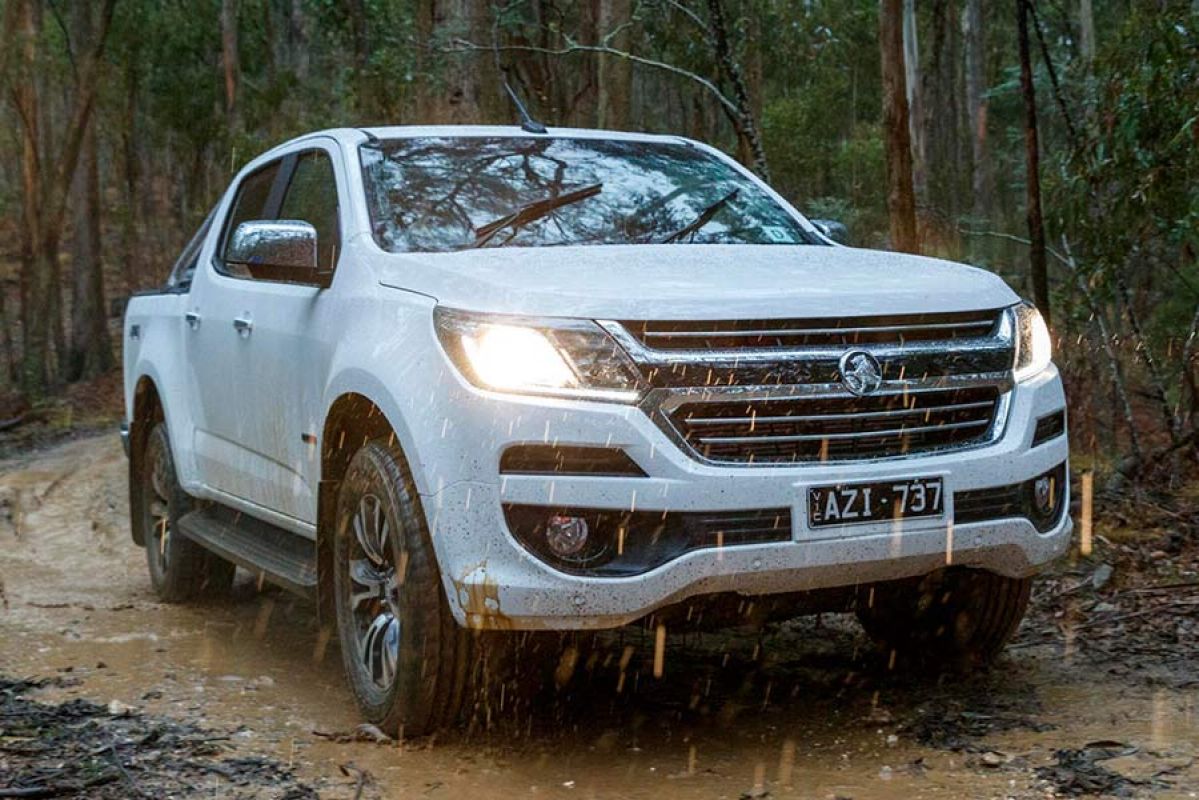 Front side view of a white Holden Colorado 2019 car driving in the rain on a muddy road through the forest