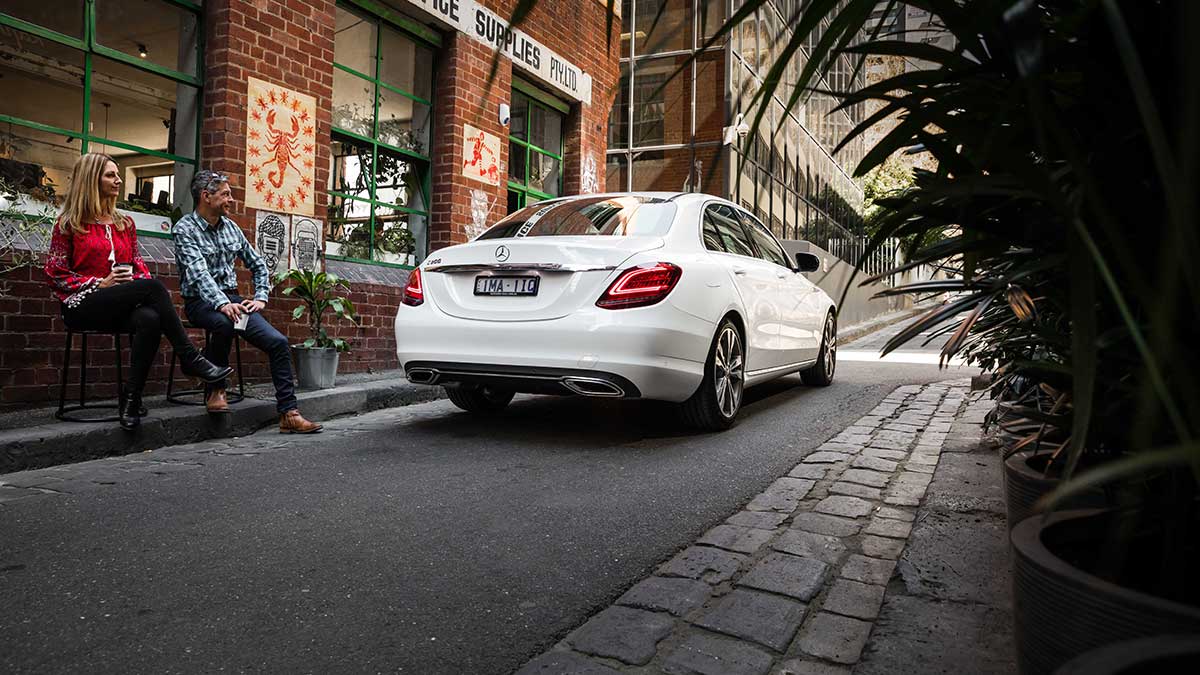 Rear side view of a white Mercedes-Benz C 200 parked in an alley next to a cafe with a man and woman sitting outside