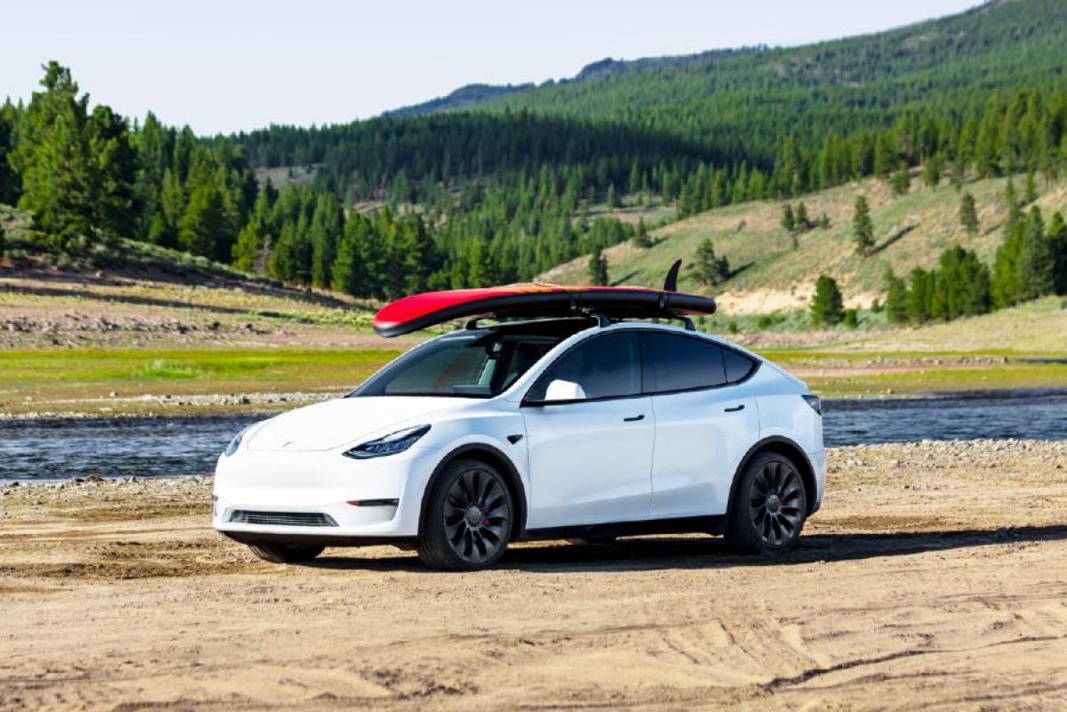 A white Tesla Model Y with a surfboard on the roof parked next to a river in front of wooded hills