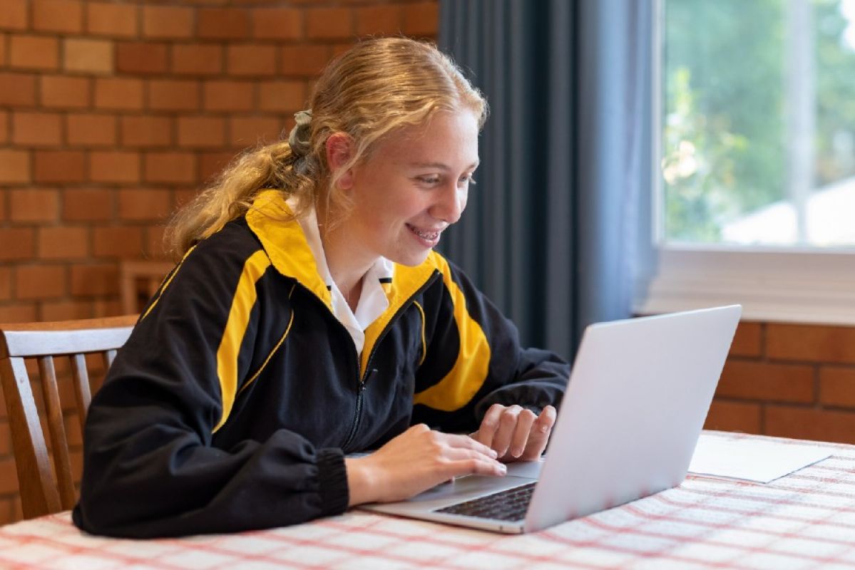 A teenage girl sitting at a table and smiling as she studies on her laptop