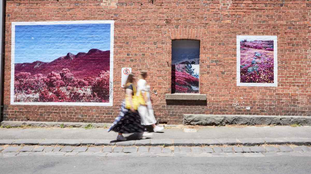 Two women walking past a brick wall with photo artworks on it