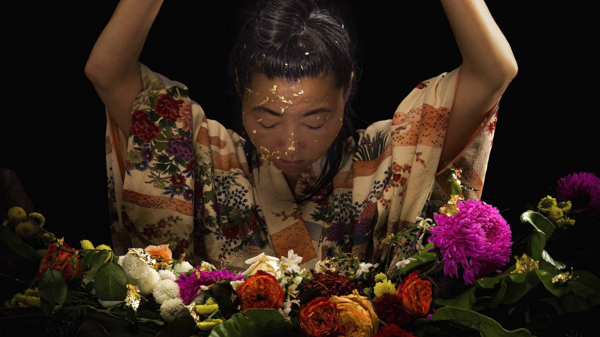 Artist Hiromi Tango with gold leaf on their face. They have their arms raised and their head bowed. In front of them are garlands of colourful flowers