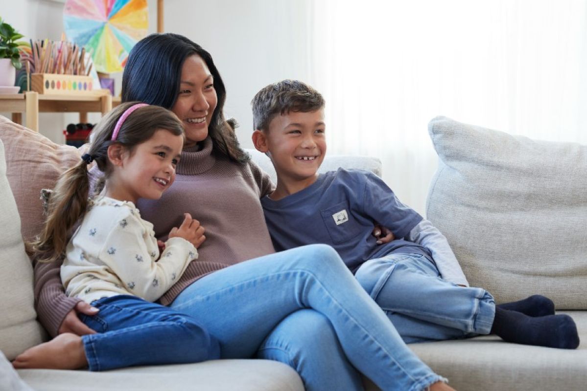 mother, daughter and son cuddling on couch smiling and looking at TV offscreen