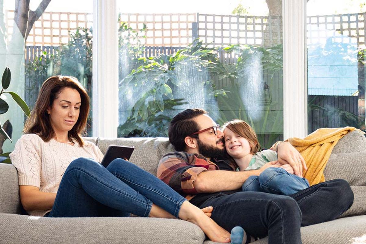 Family on couch in sun-filled loungeroom with wife looking at phone and dad cuddling young child 