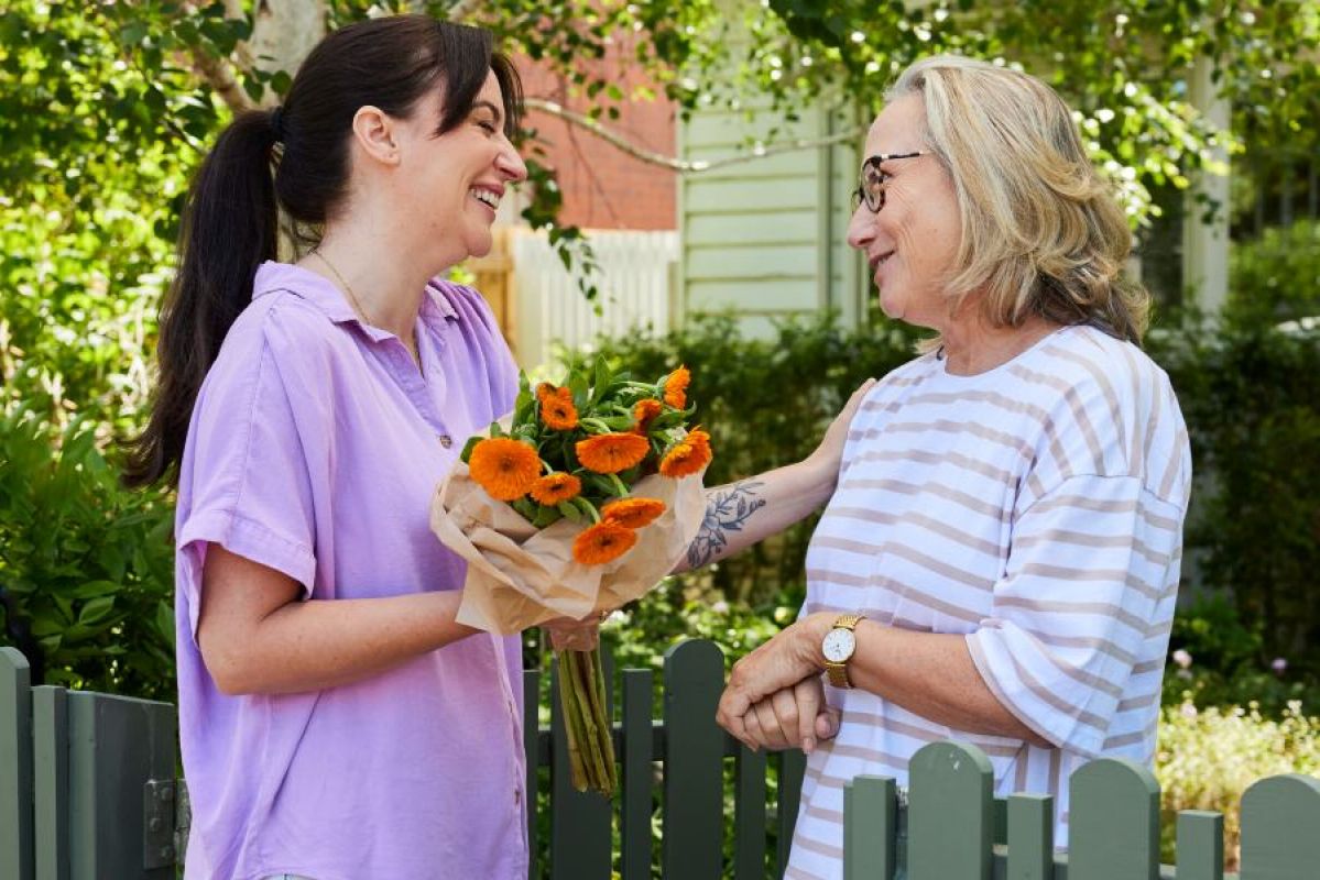 Two women, one young with long dark hair in a ponytail and one middle-age with greying blonde hair stand outside under a tree. The younger woman has brought bright orange flowers as a gift and both women are smiling