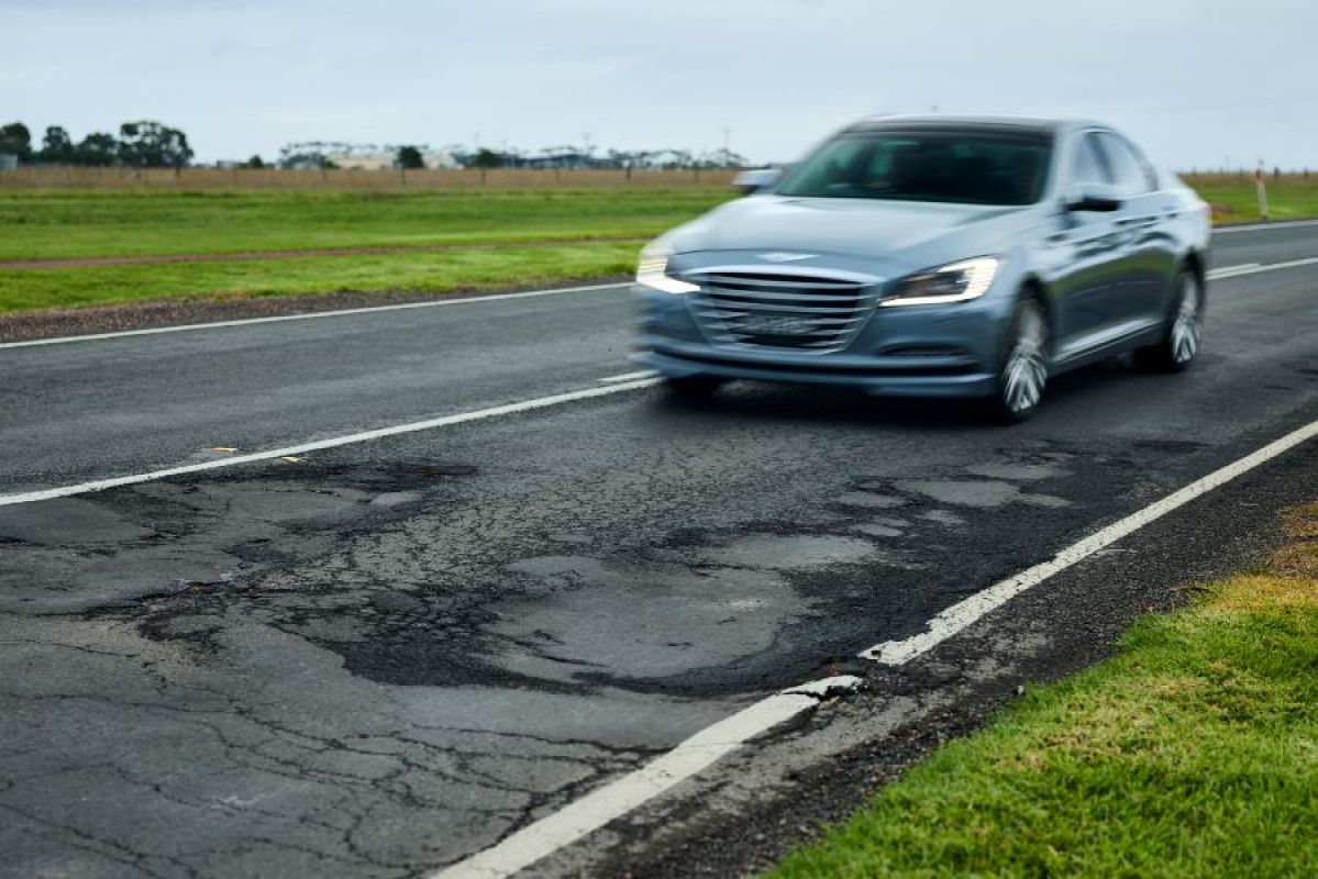 A highly weathered section of road on the Hamilton Highway. A grey sedan drives on it, about to go over a sunken, cracked part of the road surface