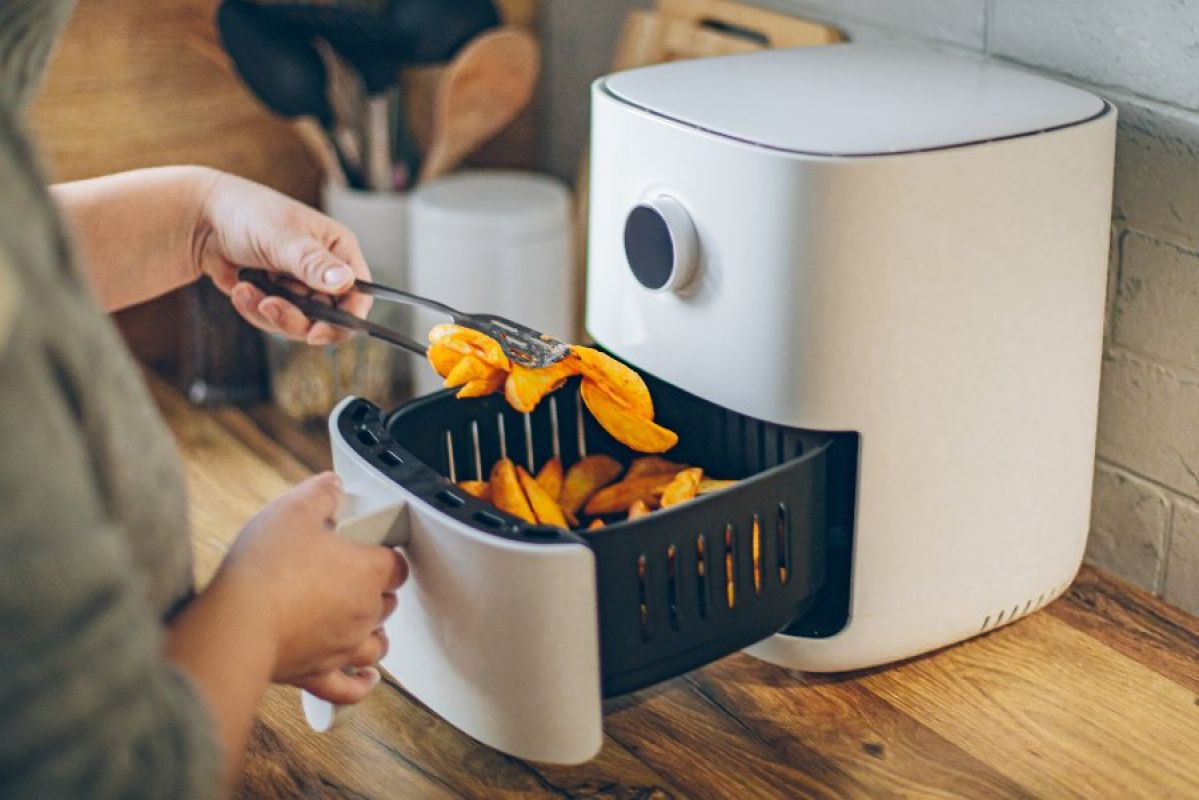 A person lifting potato wedges out of a white air fryer on a rustic kitchen bench