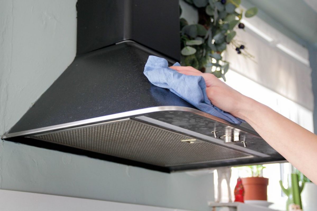 A person wiping down a dusty stainless steel canopy-style rangehood with a blue microfibre cloth
