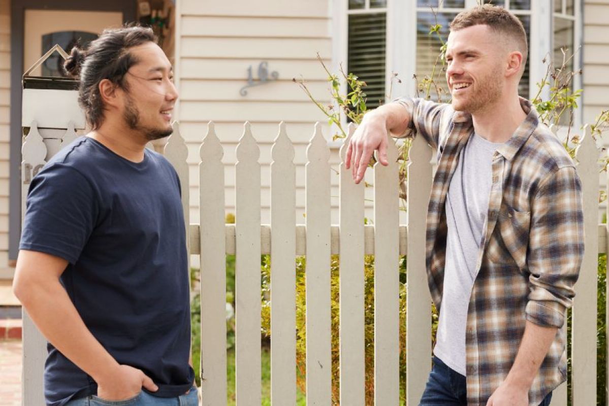 Two men leaning on a picket fence outside a weatherboard house. 