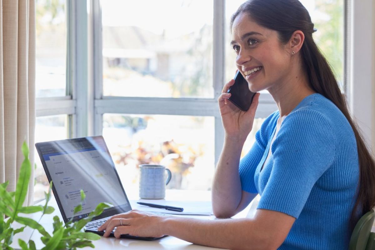 Young woman with long hair and a blue shirt smiling as she speaks on the phone to someone while also using a laptop