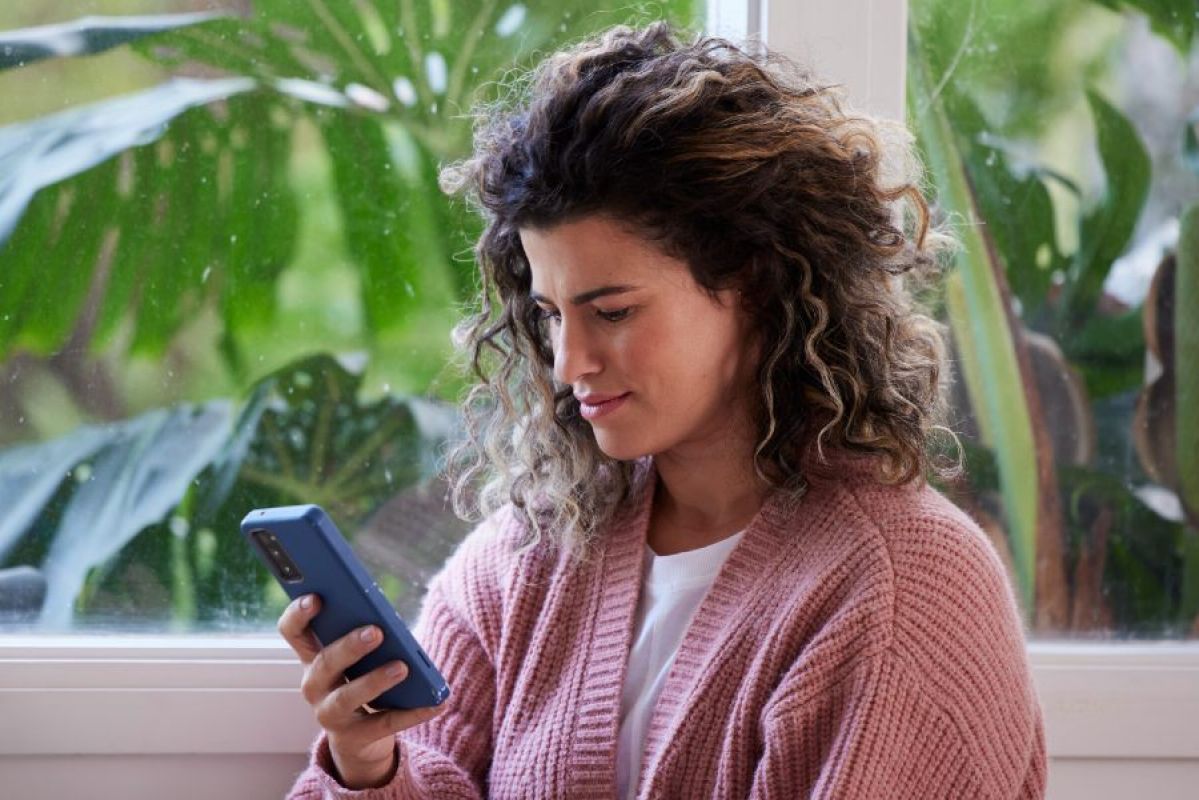 young woman smiling slightly at her smartphone as she sits near a window
