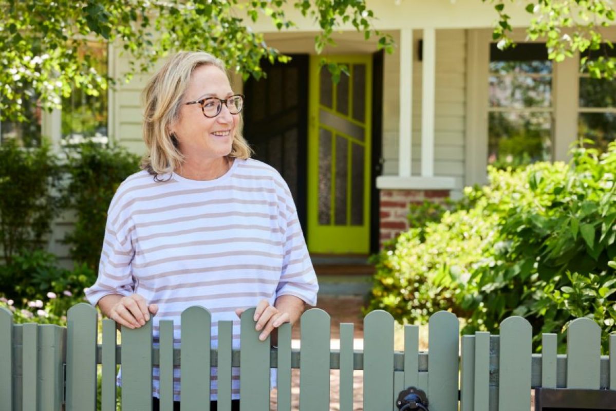 A middle-aged woman smiling as she leans on her front garden fence