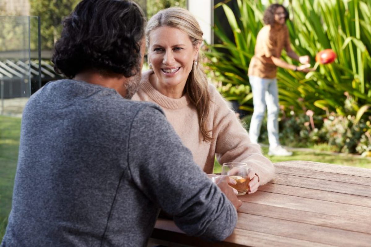 A couple chat happily at a wooden table in their backyard while their two grown children toss a footy to each other in the background