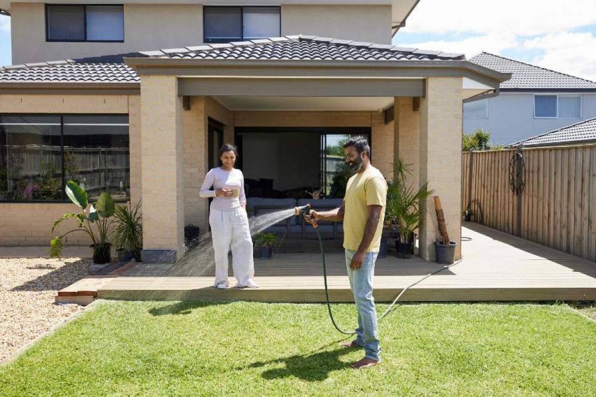 A couple stand in the backyard of a modern house. The man smiles as he waters the lawn and the woman smiles as she watches him from the deck. It is a sunny day.