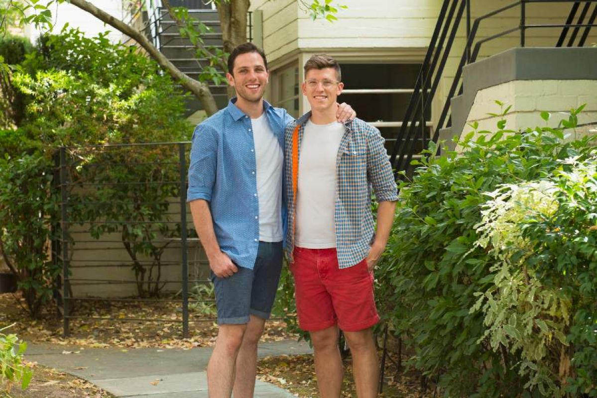 A couple smile in a leafy apartment courtyard. One man rests his hand lovingly on the other man's shoulder
