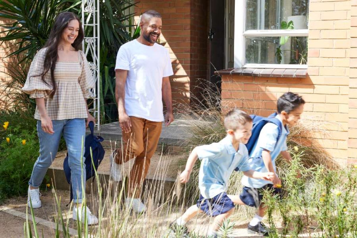 A young family - a man, a woman and two young children in school uniform, smile as they walk outside their brick house on a sunny day