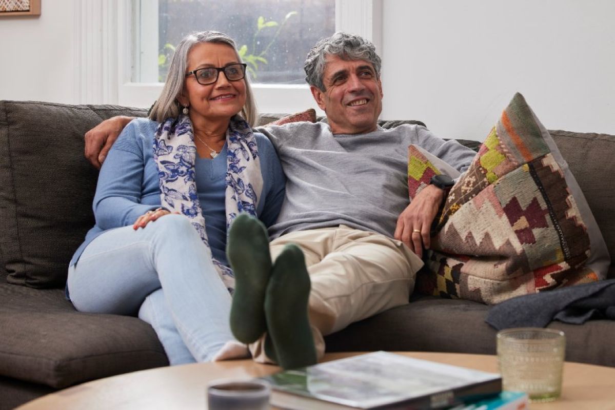 An older couple, a man and a woman, sitting comfortably together on a plush couch. The man rests his feet on a coffee table