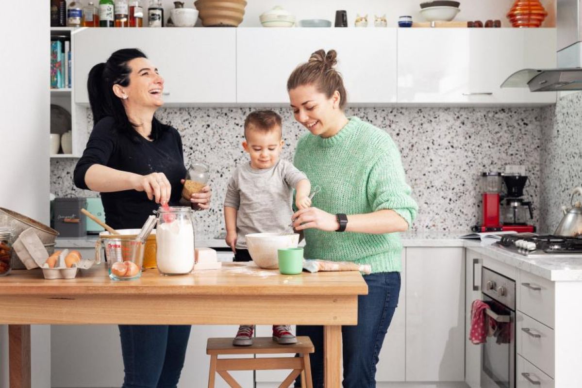 A couple smile and laugh in their modern kitchen while cooking something with their toddler