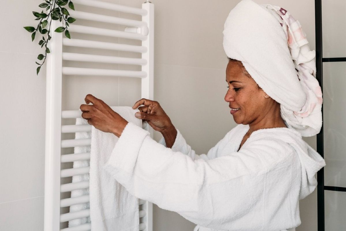 A woman wearing a white bathrobe and towel around her hair hangs a fluffy white towel on a white heated towel rail in her bathroom