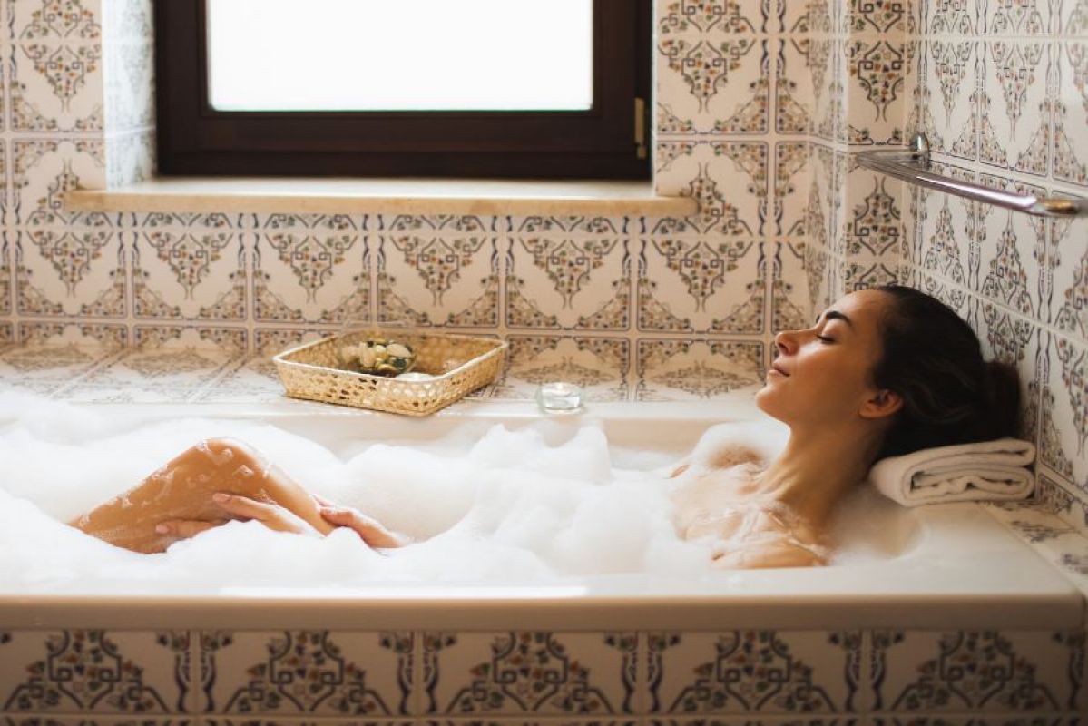 A woman enjoying a bubble bath in an ornately tiled bathroom. She is resting her head on a folded pillow and tea lights are on the bath ledge