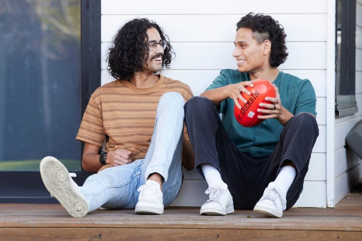Two young people sitting outside next to a white weatherboard house. They smile at each other and one holds an Aussie Rules  football