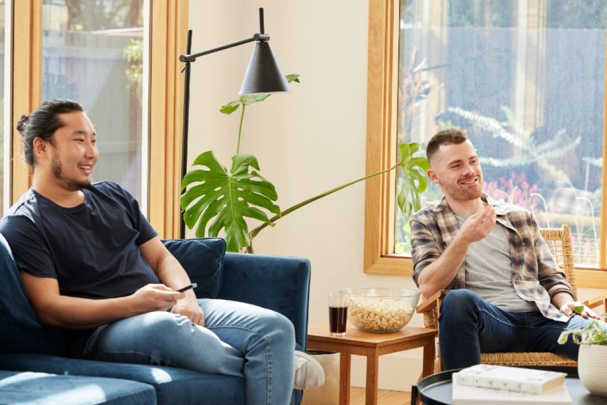 two young men in their living room smiling and watching an offscreen TV
