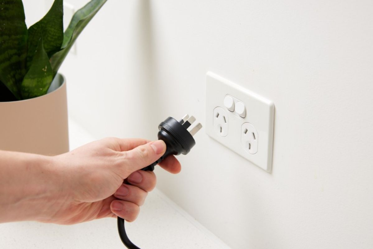 A person going to plug a black power cord into a power point in the wall