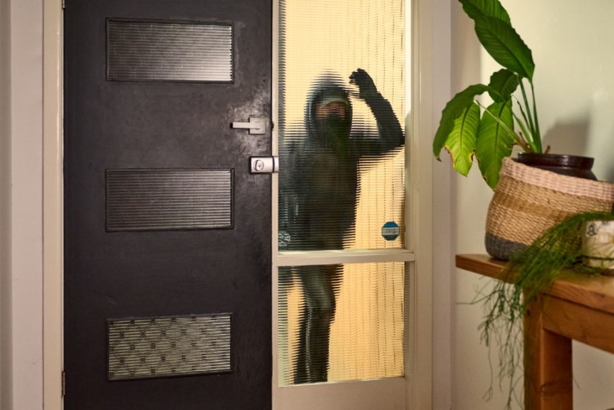 burglar in dark clothes and mask looking through front window into house