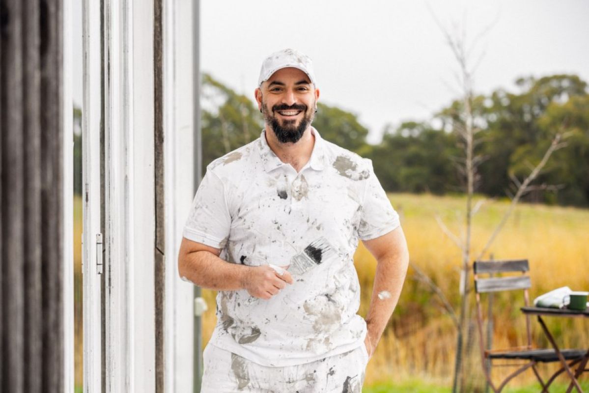 man with paint-covered clothes holding a paintbrush and smiling outside a house
