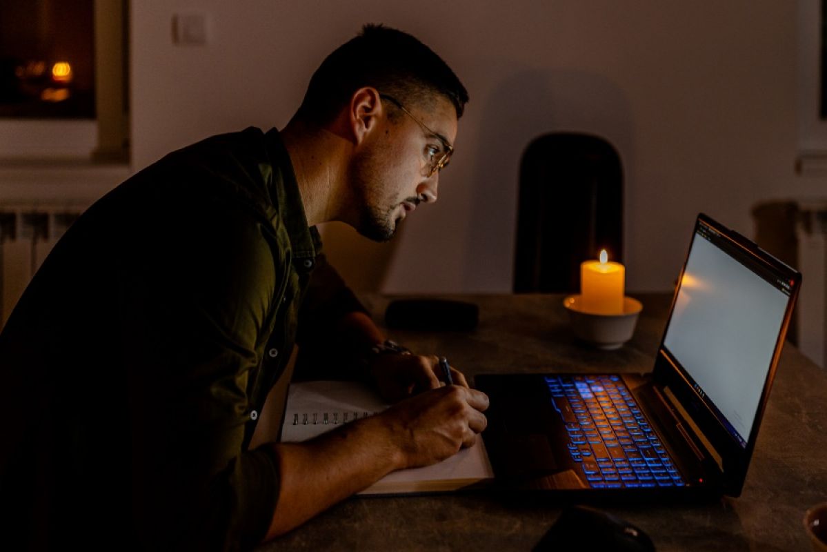 young man taking notes from laptop in a blackout with candles lit nearby
