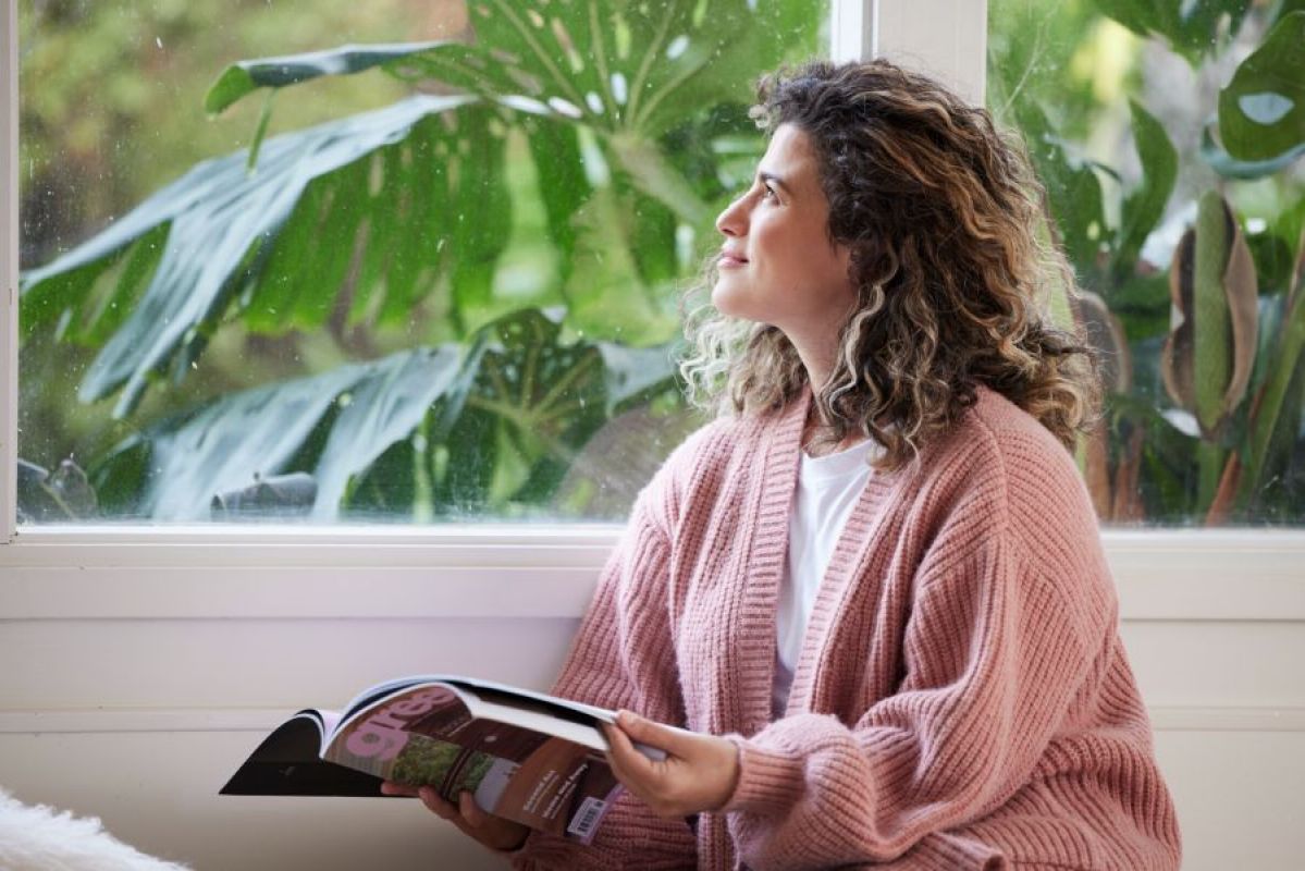 woman in pink cardigan holding a magazine and looking out a window