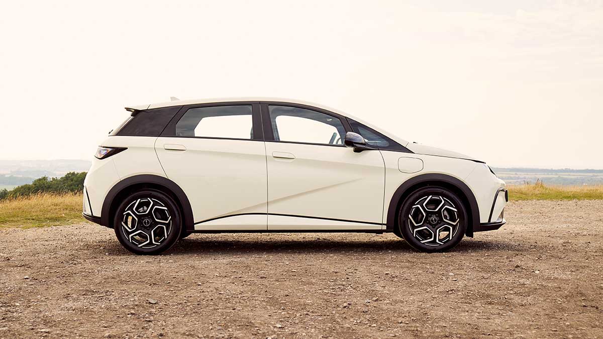 Side profile of white BYD Dolphin electric hatch in carpark with rolling hills behind. 