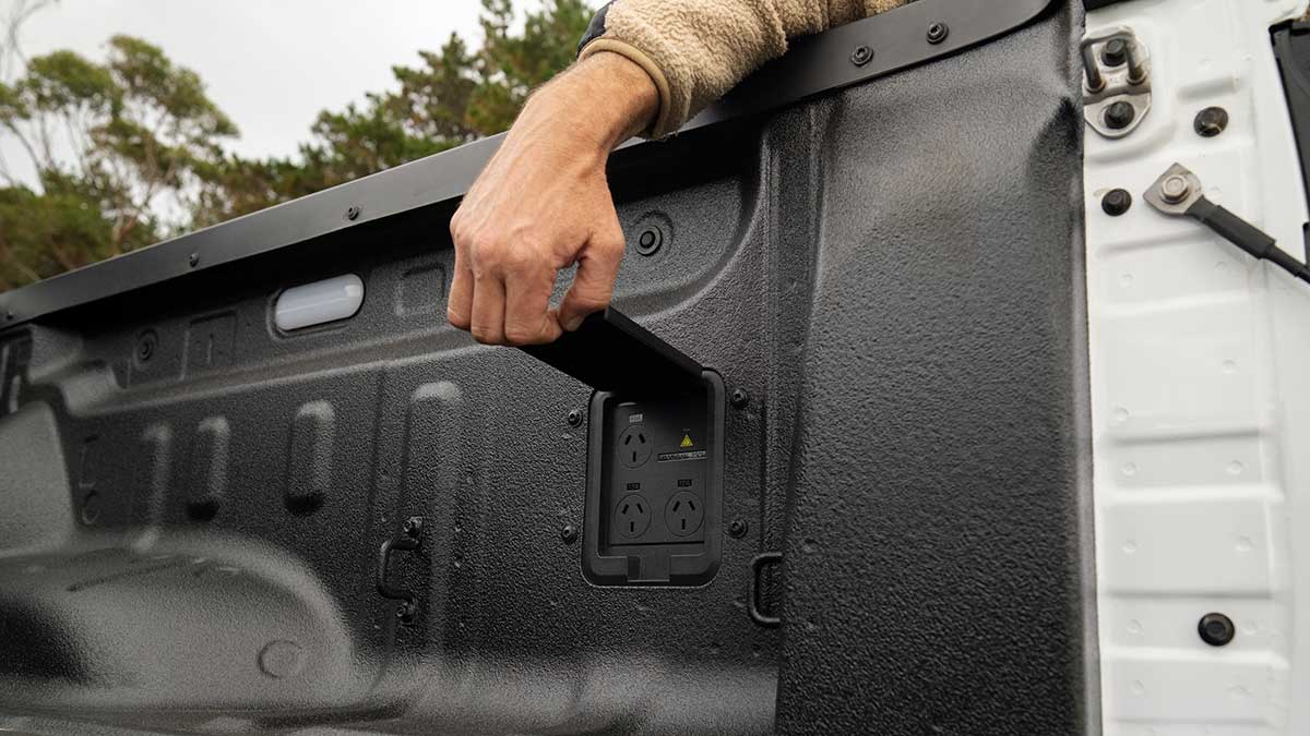 Hand reaching into load bay of white BYD Shark 6 ute, opening power V2L flap to show three power points