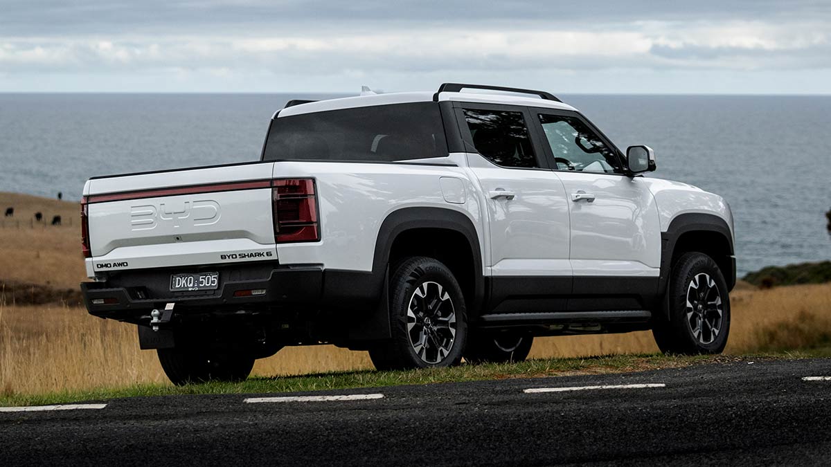 Rear quarter view of white BYD Shark 6 Premium ute parked on side of country road with farmland and ocean in background