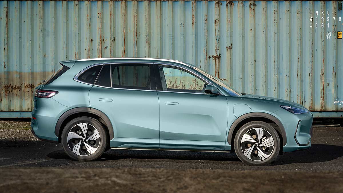 Side-on view of grey-blue Geely EX5 Inspire medium SUV in front of rusty shipping container