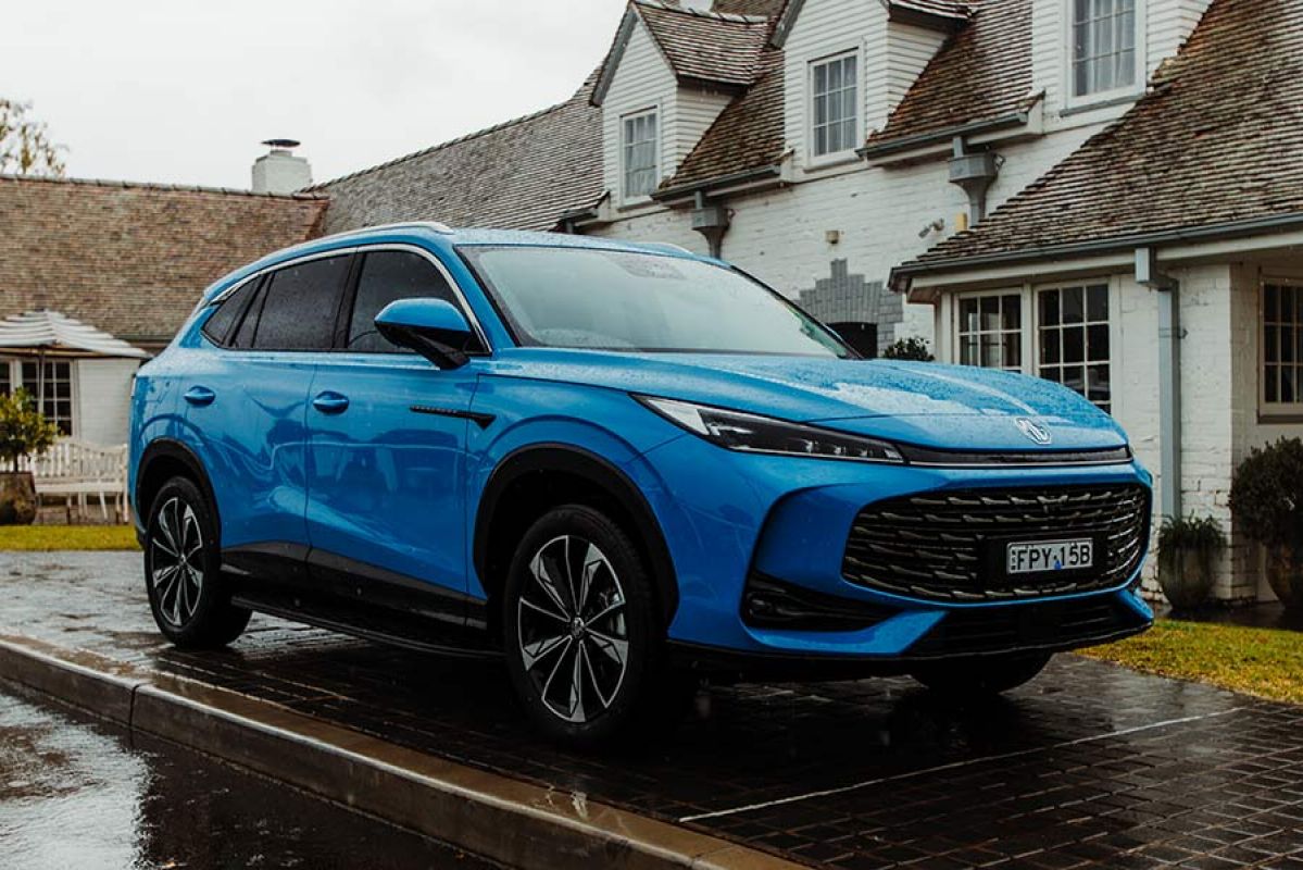 Blue MG HS medium-size hybrid SUV on wet pavement in front of white-brick heritage house.