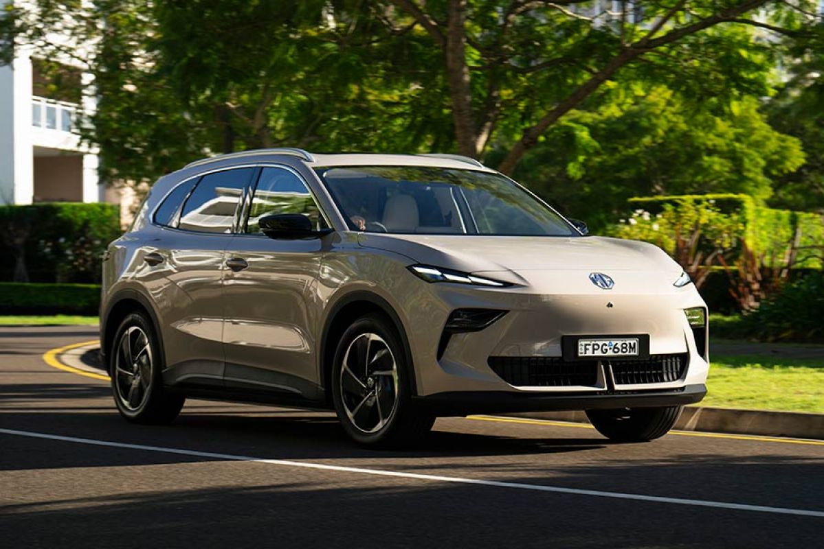 Champagne gold metallic MG S5 EV small SUV in leafy green suburban street with white house and hedgerows behind.
