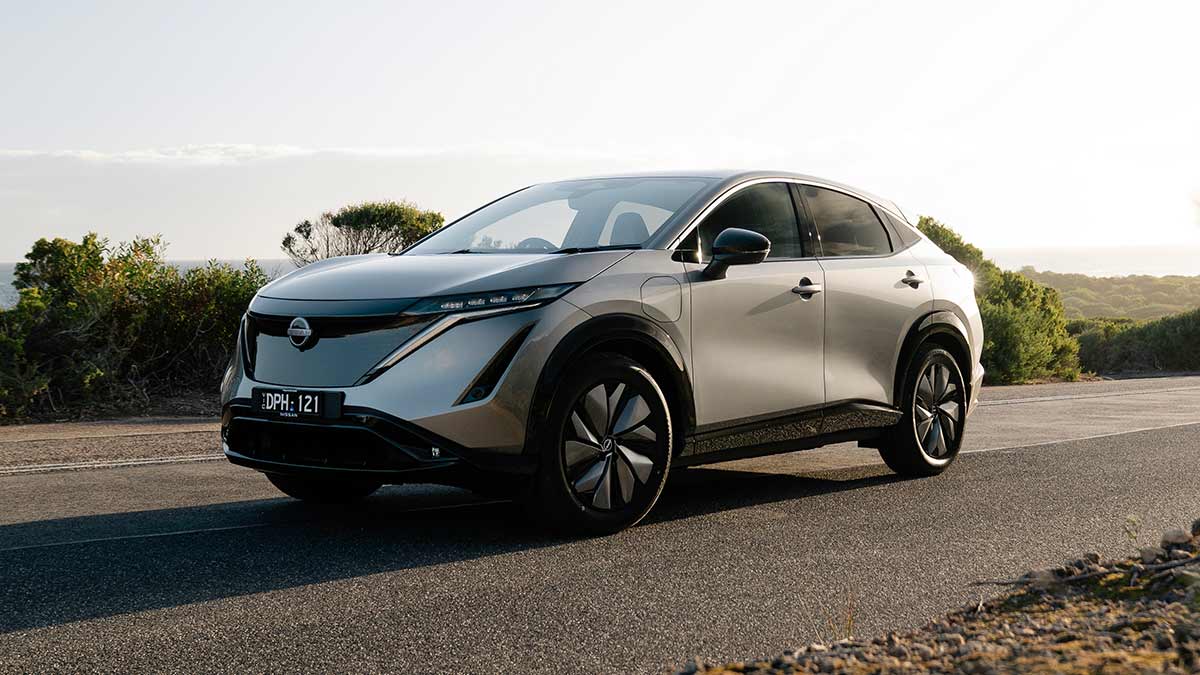 Grey Nissan Ariya electric SUV parked on side of tree-lined coastal road with ocean in background.