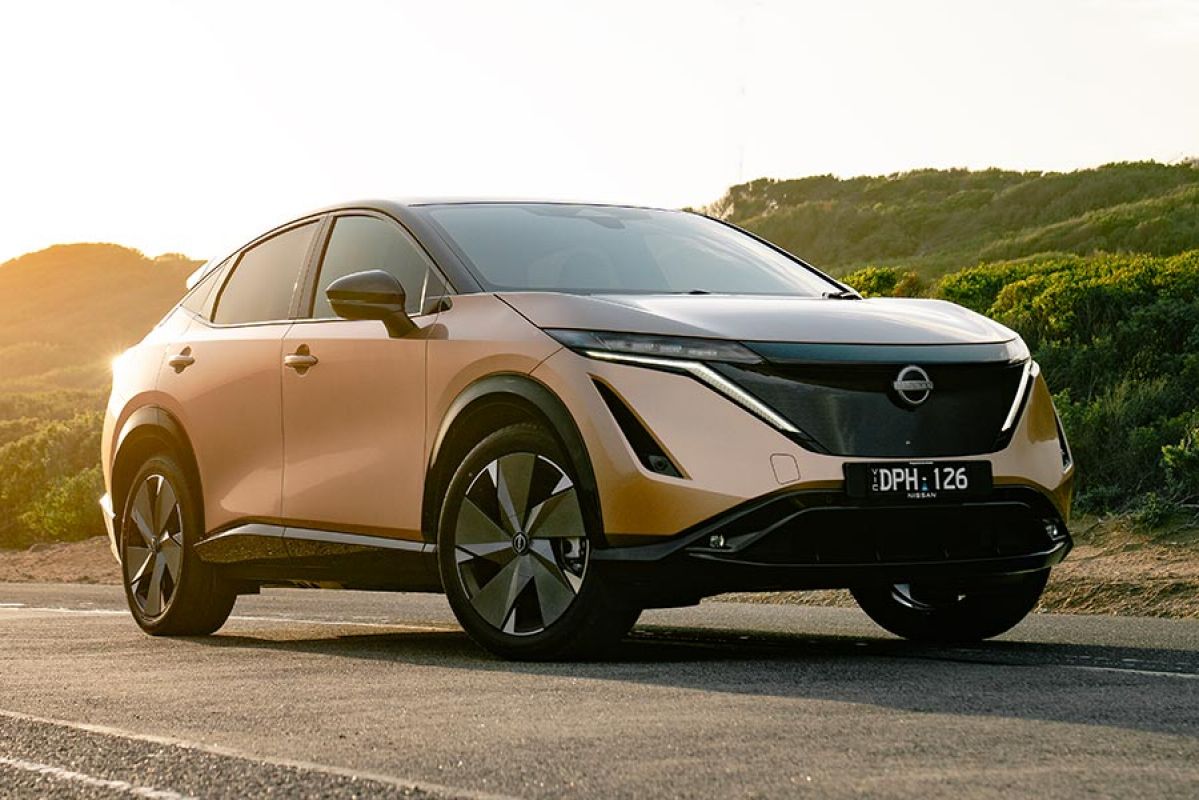 Copper-coloured Nissan Ariya Evolve electric SUV parked on side of coastal road with sun lighting up rear of car.