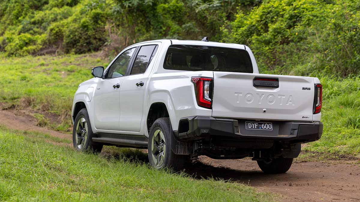 Rear view of white Toyota HiLux SR5 EV dual-cab ute on off-road track in open grassy clearing.