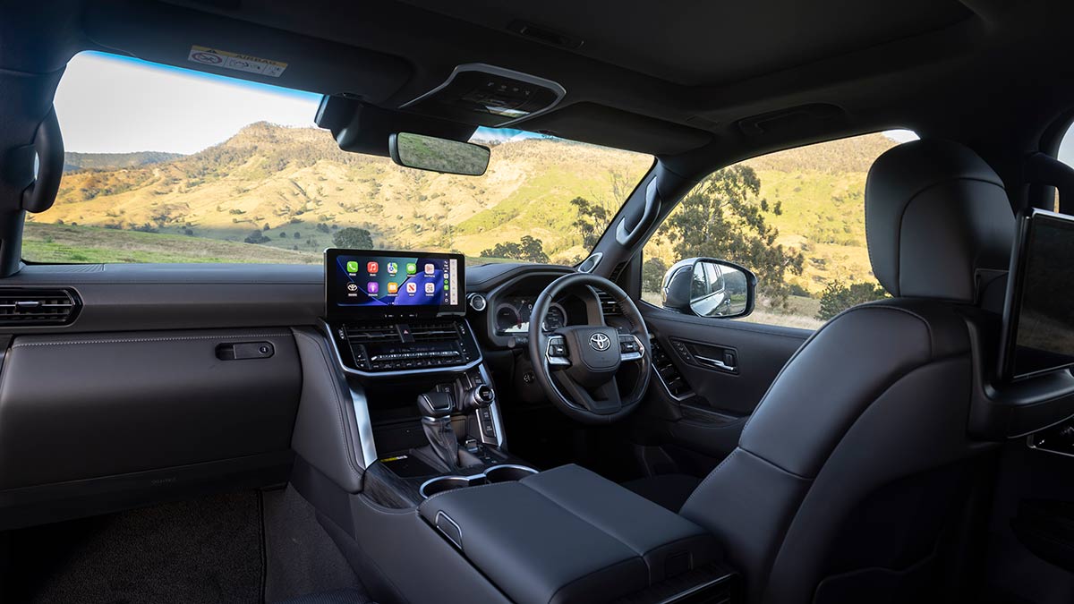 Cockpit area of Toyota LandCruiser Hybrid Sahara ZX with dark leather-accented trim, broad switchgear and dual digital screens.