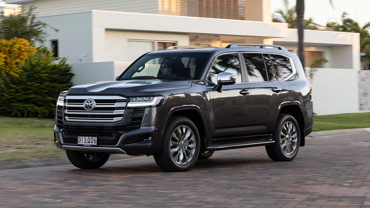 Dark grey metallic Toyota LandCruiser Sahara ZX Hybrid SUV on suburban street passing white modern house.