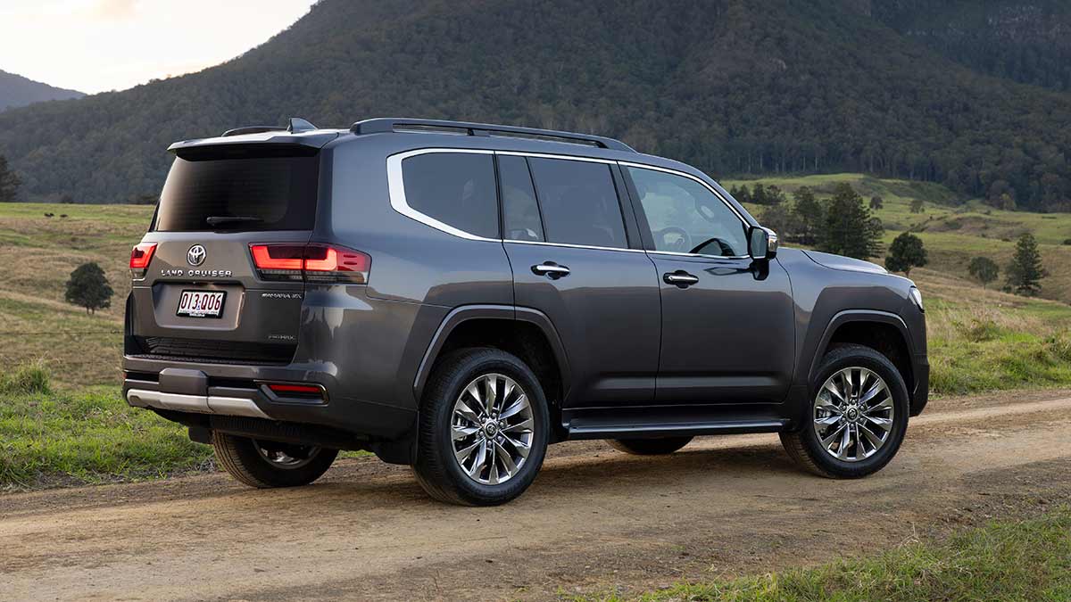 Rear view of dark grey metallic Toyota LandCruiser Sahara ZX Hybrid SUV on dirt road in alpine country.