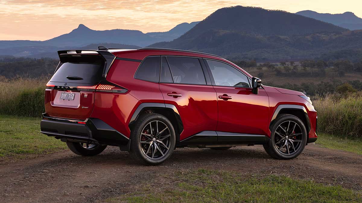 Rear three-quarter shot of red Toyota RAV4 GR Sport medium SUV parked across dirt track at base of mountain range at dusk.