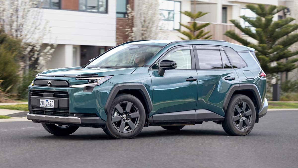 Metallic green Toyota RAV4 Edge SUV driving on suburban street in front of white townhouses.
