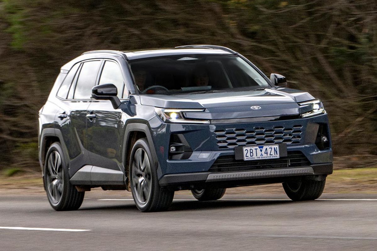 Metallic blue Toyota RAV4 SUV driving on country road with green verge and thick native scrub behind.